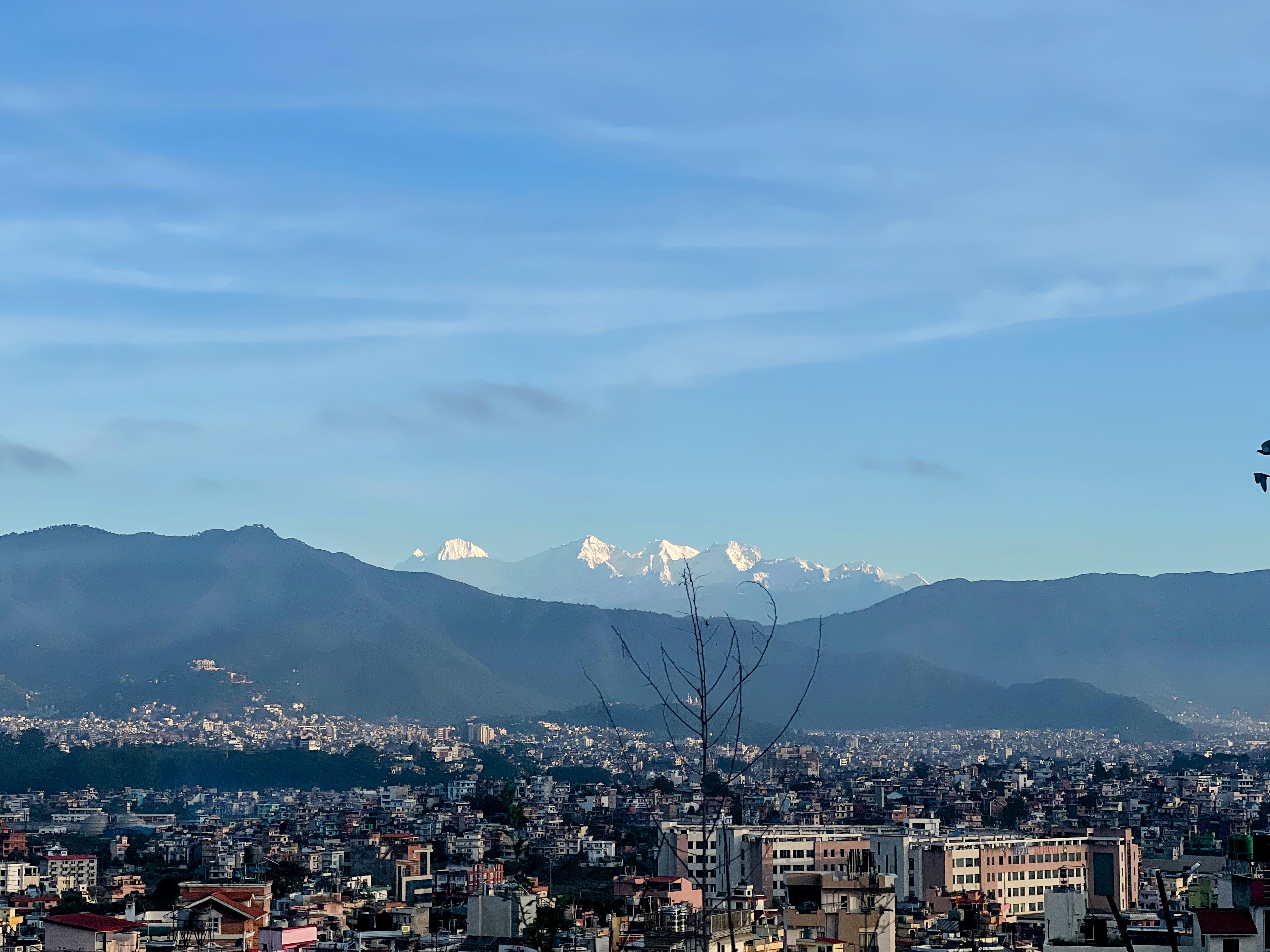 A wide cityscape view showing a dense urban area in the foreground and snow-capped mountains rising in the distance under a clear blue sky.