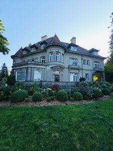 A wide view of the back side of Pittock Mansion, Portland, taken in the evening. It shows the classic stone architecture, red roof, and well-kept garden.