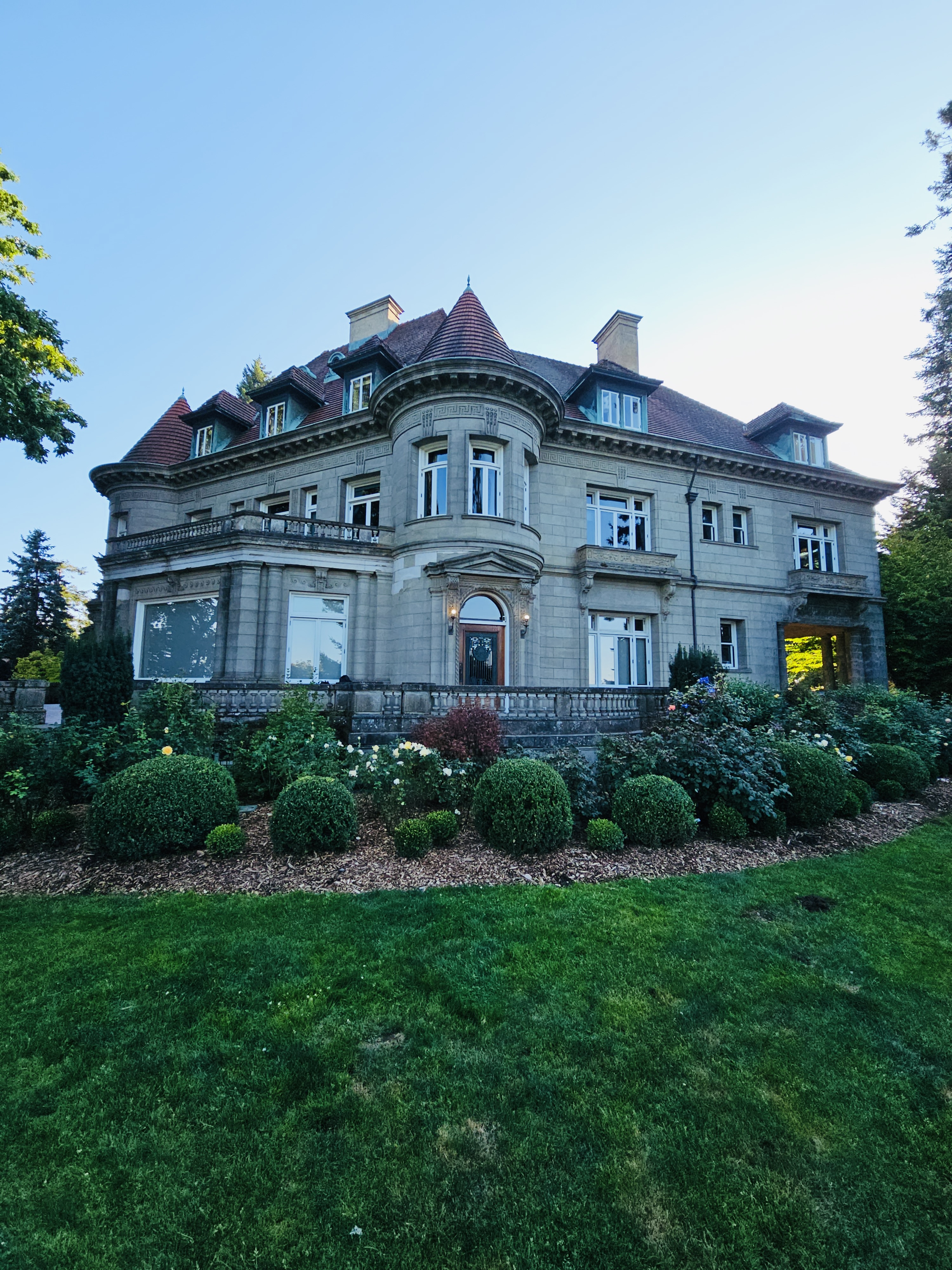 A wide view of the back side of Pittock Mansion, Portland, taken in the evening. It shows the classic stone architecture, red roof, and well-kept garden.