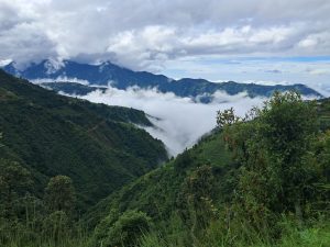 Lush green hill and mountain vista under a cloudy sky. A thick layer of clouds fills the valley below, partially obscuring the landscape.