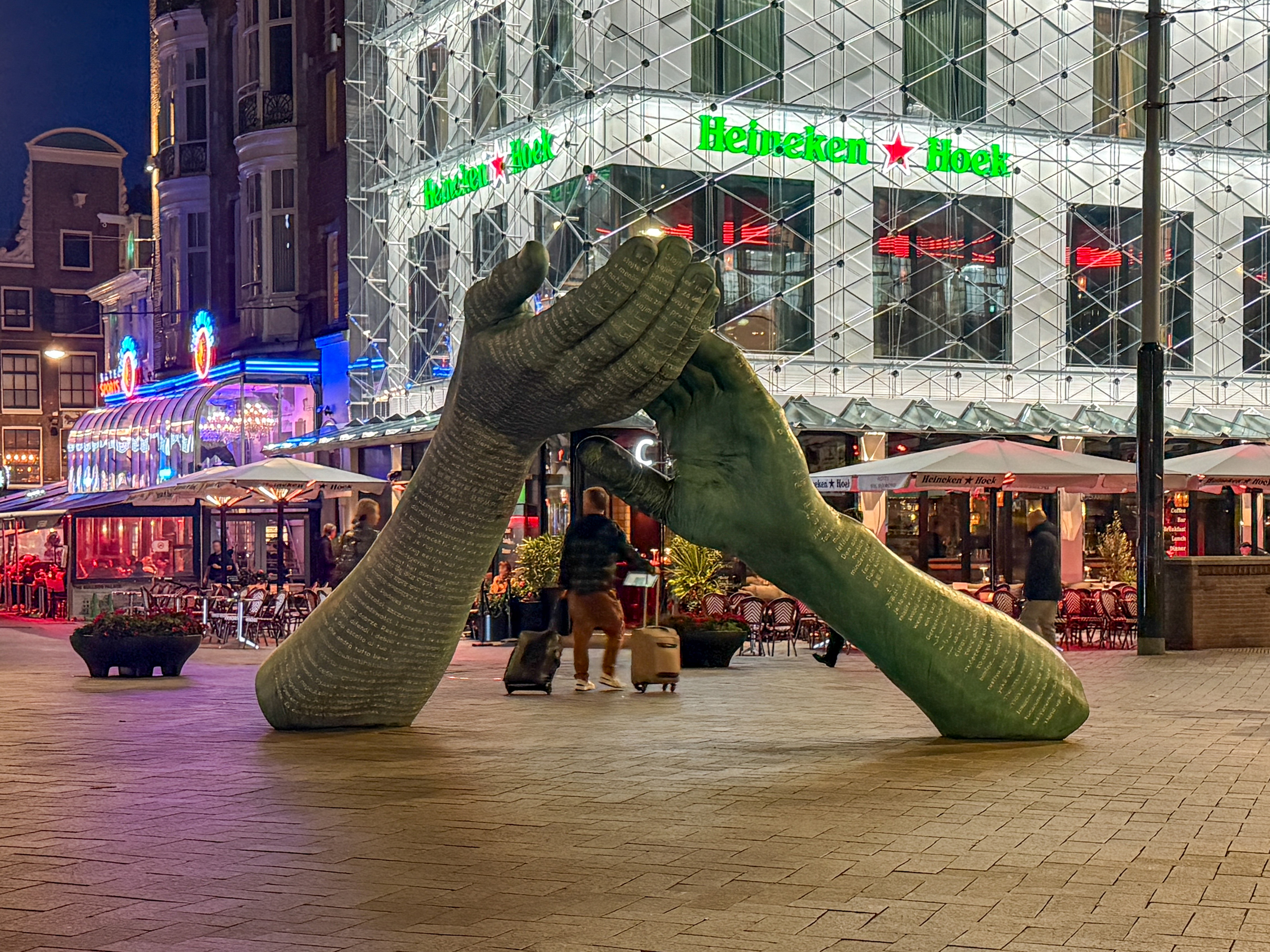 A large outdoor sculpture of two oversized hands clasping each other stands in a busy plaza at night. Behind it, the Heineken Hoek building glows with bright lights and neon signs, while people walk and sit at café tables under umbrellas along the lively street.