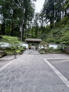 A traditional wooden Japanese gate stands at the entrance of the Japanese Garden, Portland, framed by stone paths and tall forest trees in the background. 