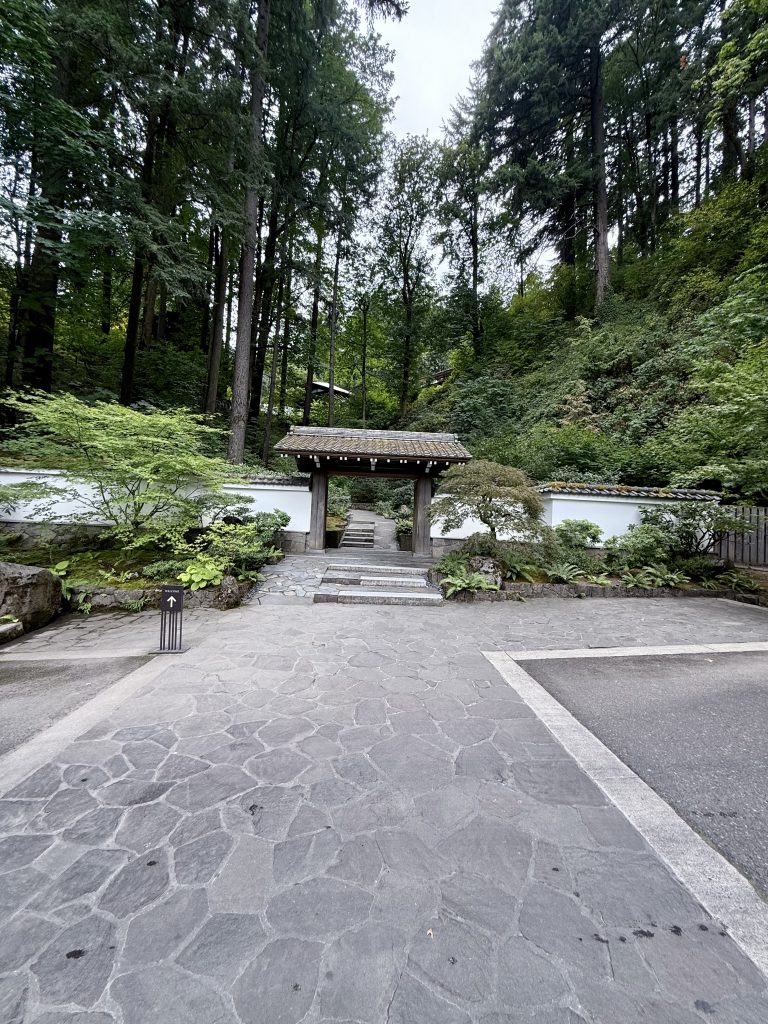 A traditional wooden Japanese gate stands at the entrance of the Japanese Garden, Portland, framed by stone paths and tall forest trees in the background.