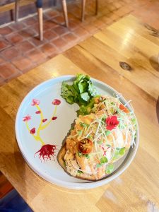 A single piece of open sandwich artisan bread topped with a creamy white sauce, egg benedict, fresh greens, red cherry tomato halves, and delicate red flowers. The food is arranged on a light blue plate with a decorative red and yellow plant-like design painted on the side. 