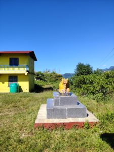 A yellow house with a red roof sits amid greenery under a bright blue sky, with a Nandi statue on a stone platform in the foreground.