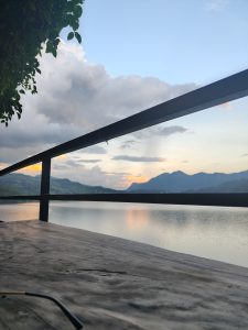 A tranquil lakeside at sunset, with calm waters reflecting mountains and a colorful sky, framed by a wooden table, leaves, and a railing.