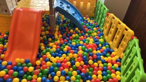 A colorful children's play area features a ball pit filled with plastic balls and two small slides, all enclosed by a bright plastic fence.