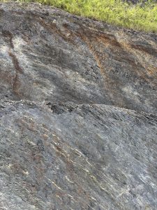A close-up view of textured rock layers, showcasing a mix of dark gray and black hues with subtle streaks of brown and gold. 