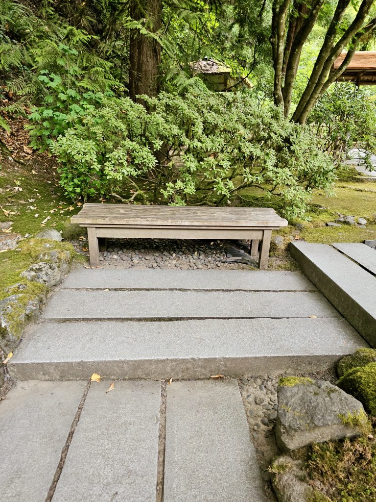 A simple wooden bench placed beside stone steps surrounded by dense greenery, offering a quiet resting spot in the Portland Japanese Garden.