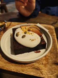 A brownie with cream, fruit, seeds, and zest on a plate, with a hand holding a spoon nearby.