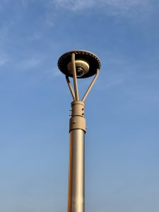 Surveillance camera on a modern light pole against a blue sky in downtown Portland. The pole features a clean, metallic finish and triangular support arms that hold the camera dome. 