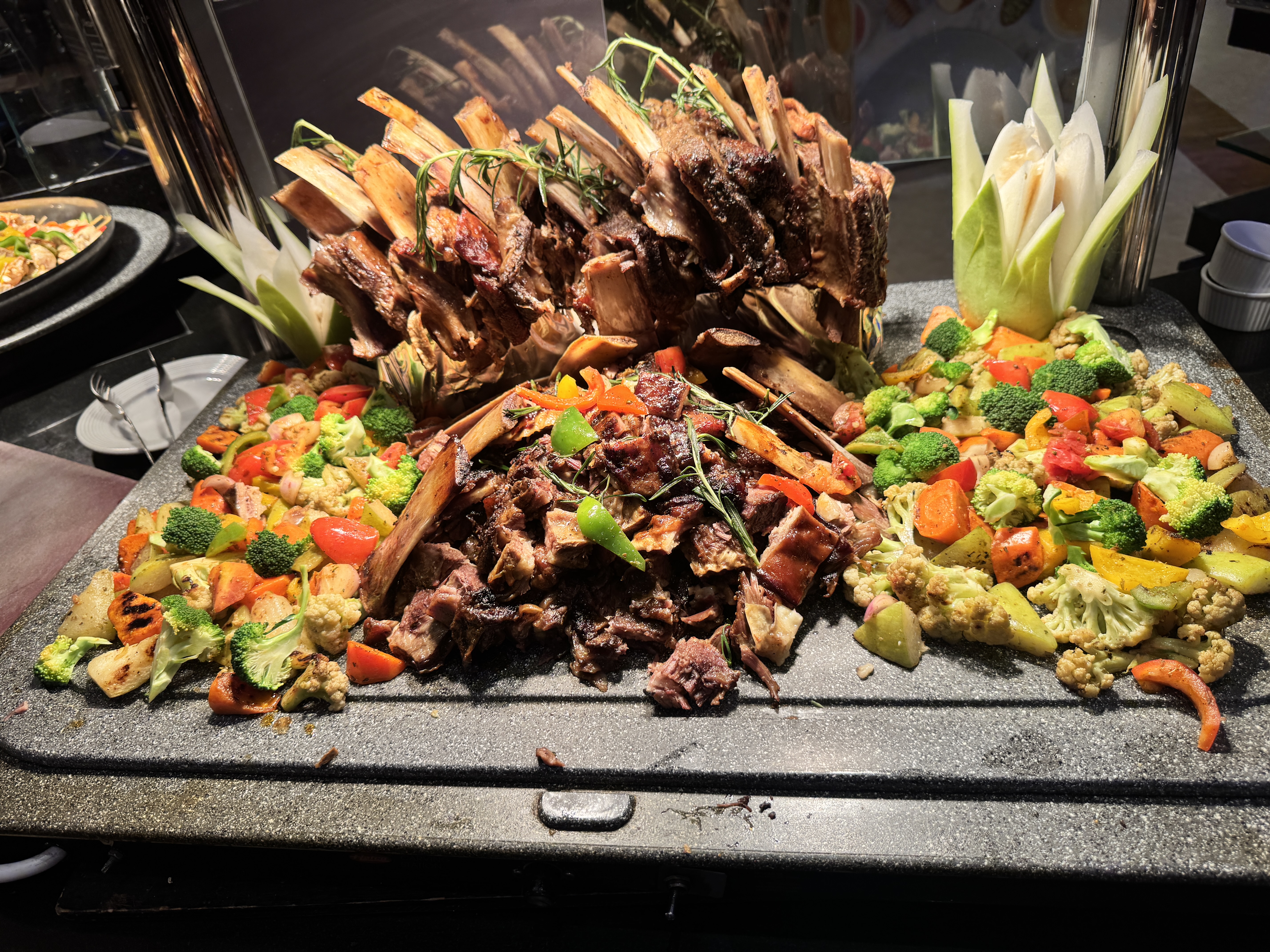 An elaborate buffet display features a large roasted rack of mutton and chopped barbecue surrounded by a colorful medley of roasted vegetables.