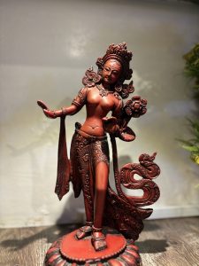 A red-colored statue of a graceful figure, possibly a deity, standing on a flower-shaped base