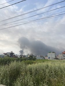 Suburban buildings with tall grass in the foreground, power lines overhead, and dark smoke rising against a cloudy sky.