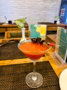 A martini glass filled with a thick orange-red cocktail is garnished with a skewer of dark, dried fruits or spices, resting on a textured placemat at a wooden table.