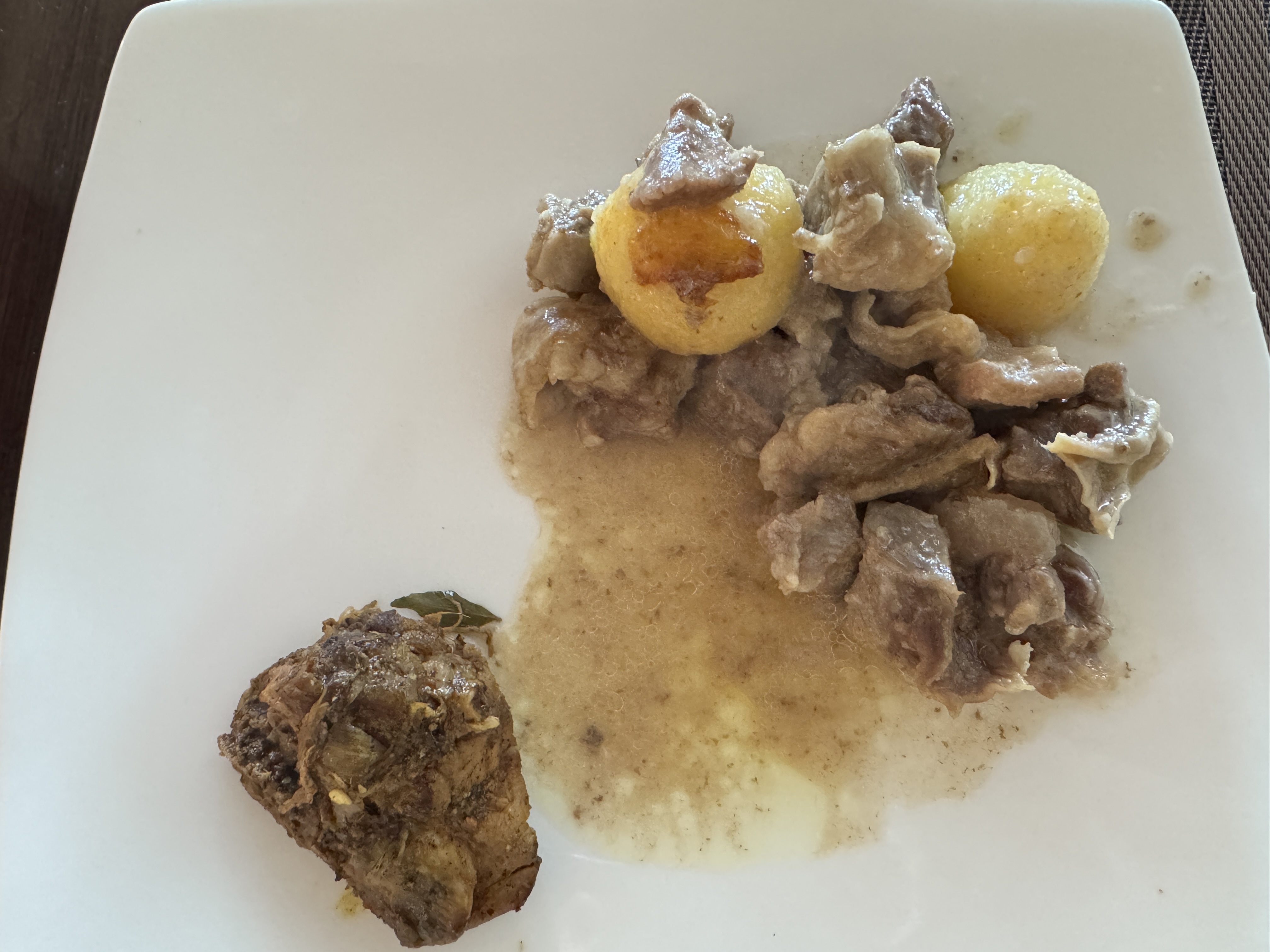 A white plate holds a small serving of stewed meat in a light gravy with two roasted potatoes and another piece of dark meat.