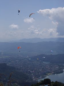 A panoramic view from a mountain overlooking a town and lake below, with several colorful paragliders soaring in the sky. 