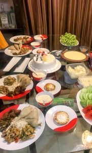 A glass table is set with a white birthday cake, several plates of savory food including momo or dumplings, fruits, and desserts.