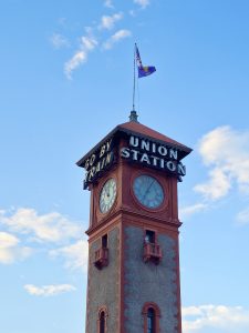The iconic Union Station clock tower, featuring the words "Go By Train" and "Union Station," stands tall against the blue sky in downtown Portland. 