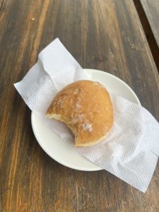 A close-up shot of a half-eaten custard filled doughnut with powdered sugar, resting on a white paper napkin on a small white plate. The plate is on a wooden table. 