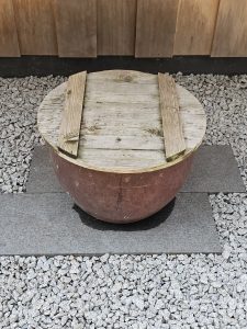 A half-buried pot with a wooden lid placed on top, sitting on gravel. Captured at Portland Japanese Garden.