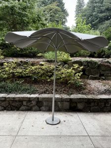 A modern grey umbrella with curved edges stands alone on a concrete path, surrounded by greenery and a stone wall in Washington Park, Portland. 