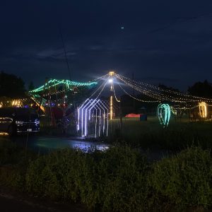 A nighttime scene featuring a playground illuminated by colorful string lights. Green and white lights form shapes including hearts and arches, creating a festive atmosphere. In the foreground, a dark-colored van is parked near the edge of the area, while a grassy patch is visible. The sky is a deep blue, indicating twilight, and some structures can be seen in the background.