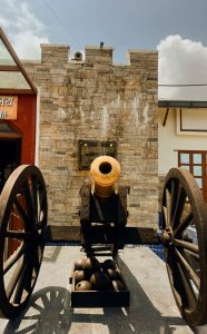 A  historic black cannon with a bright golden barrel is displayed on a platform in front of a stone wall structure resembling a turret, with its two large wooden wheels flanking the scene.