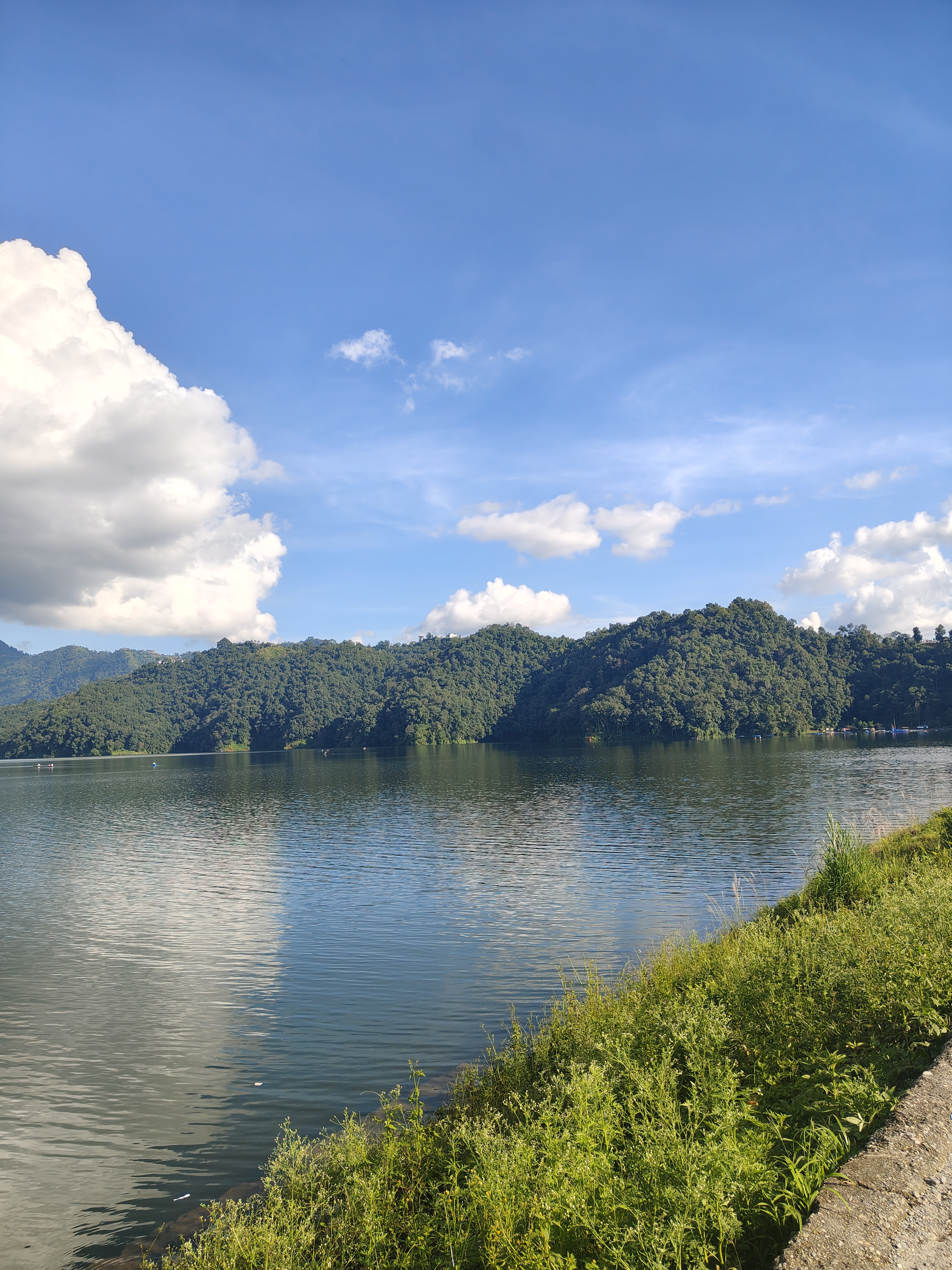 A serene Nepal landscape with a calm lake, green hills, and a blue sky with white clouds.