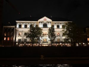 A grand, ornate building illuminated at night, its white façade glowing with architectural details highlighted by spotlights. Trees partly frame the structure, while reflections of light shimmer on the dark canal in the foreground.