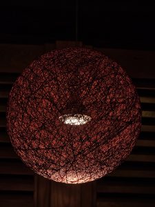 A handmade round lamp made of red threads surrounds a light bulb. The warm light glows through the threads, creating a cozy feel. Taken from downtown Portland. 