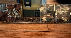 A close up picture, two glasses on the right filled with ice and clear fluid, on the left two measuring jiggers.  They're sitting on a metal panel on a wooden bar.