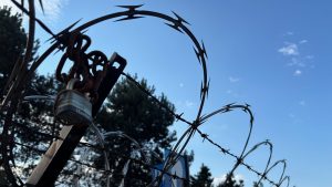Set against a clear sky, some razor wire wrapped around barbed wire.  In the foreground is a padlock.