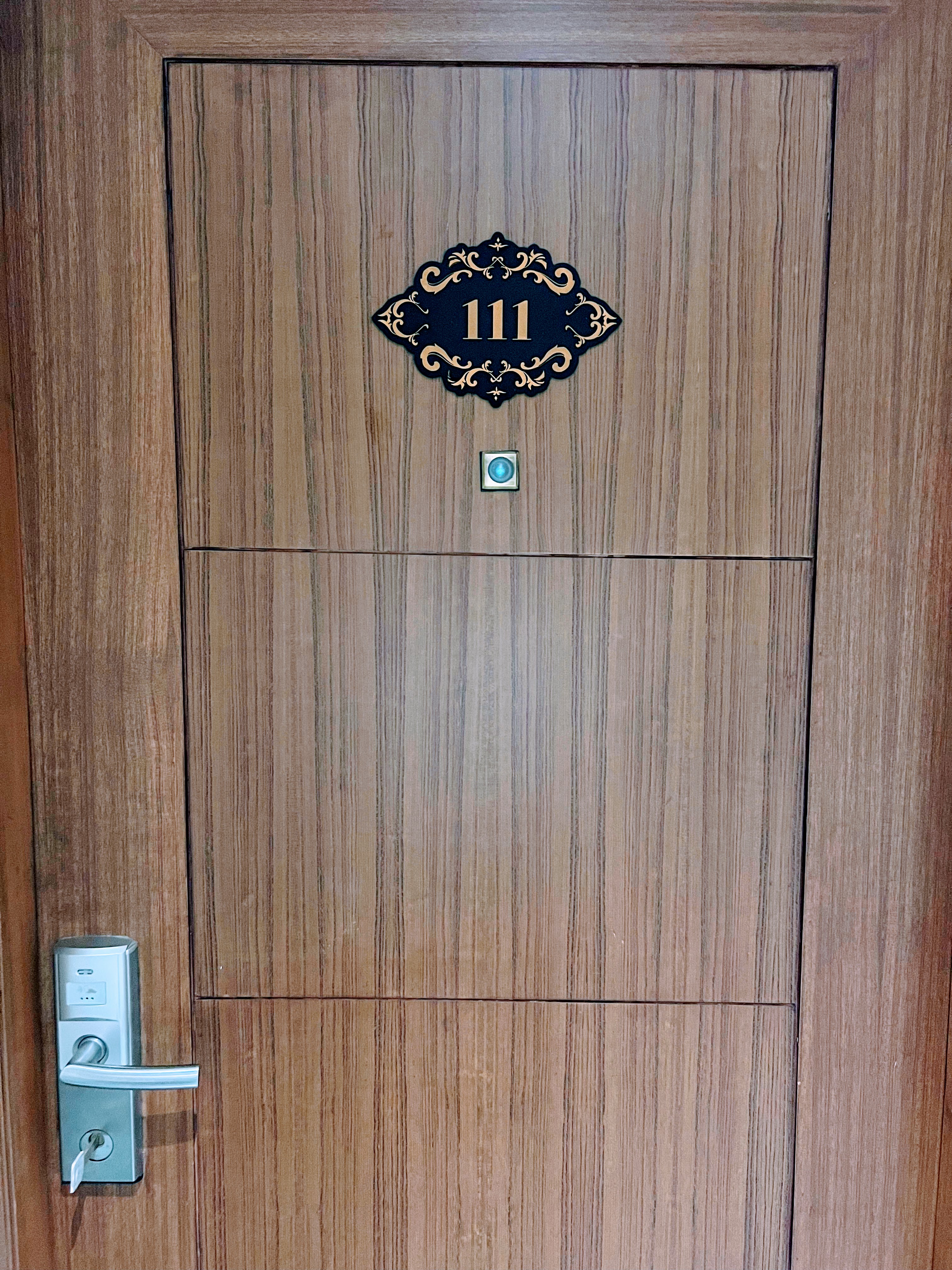 A hotel room door with a wood grain finish, gold plaque above the peephole, and a silver key card lock.