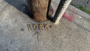 On the sidewalk at the base of a wooden pole the letters "R O E K" with stars on either side and an arrow pointing at them, painted in some kind of puffy paint.