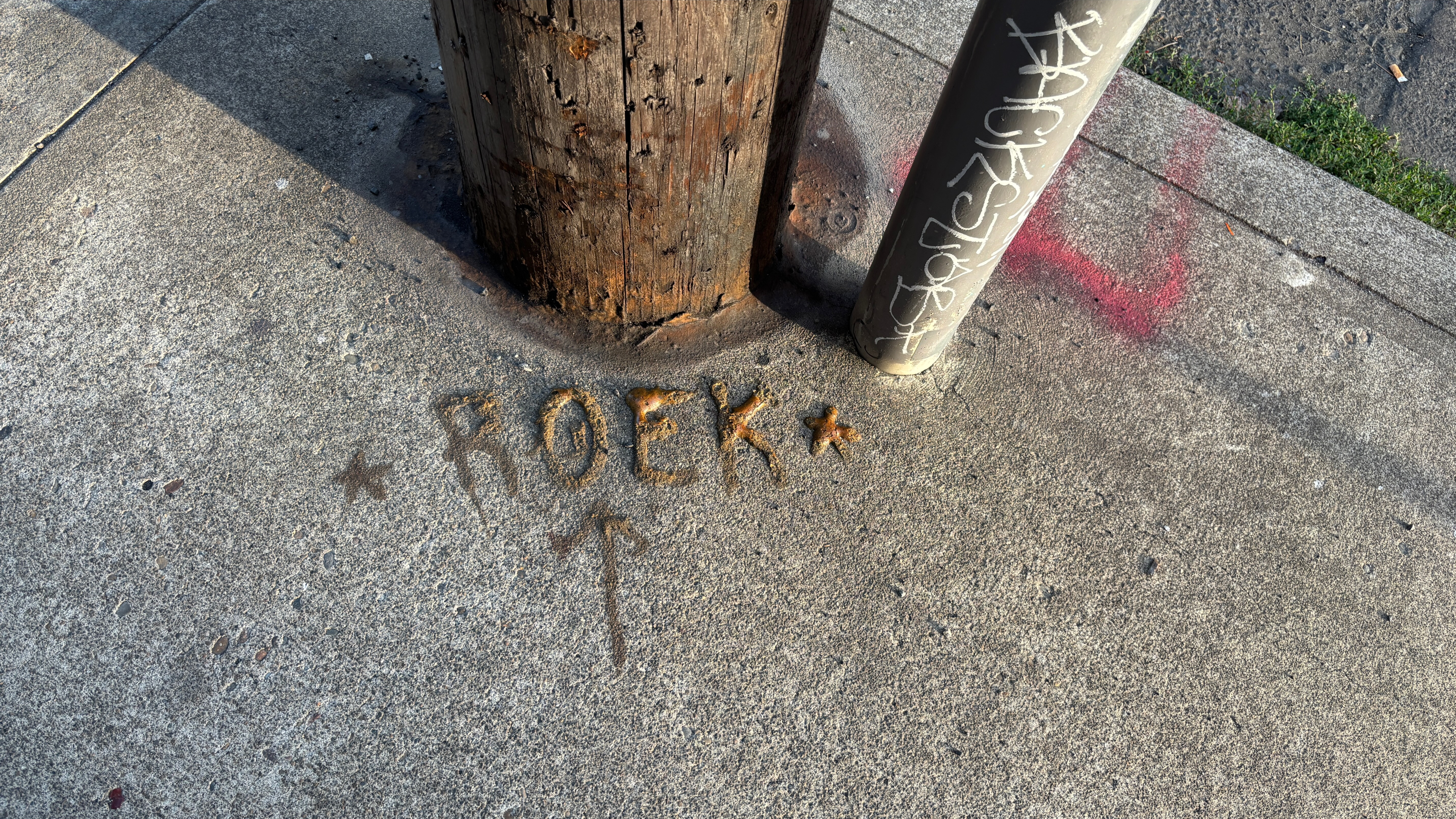 On the sidewalk at the base of a wooden pole the letters "R O E K" with stars on either side and an arrow pointing at them, painted in some kind of puffy paint.