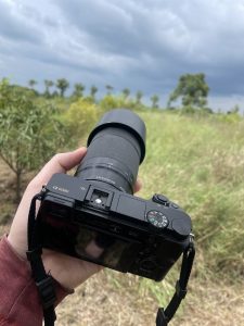 A person holding a Sony Alpha 6000 camera with a telephoto lens in an open grassy field under a cloudy sky.