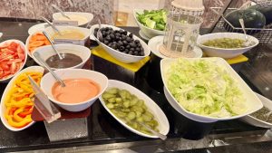 A salad bar is laid out with many white bowls containing fresh ingredients like lettuce, peppers, and olives, as well as various dressings.