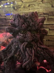 A close-up image of an ornate stone-like sculpture depicting a mythical creature with an expressive face, characterized by a gnarled beard and hair resembling tree branches.