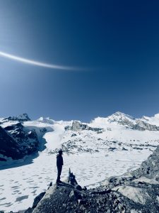 A person stands on a rocky ledge, silhouetted against a vast snowy landscape. In the background, icy glaciers and snow-covered mountains rise under a clear blue sky. 