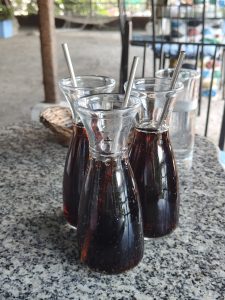 Three dark-filled glass bottles with metal straws sit on a granite table, alongside a glass of water, with greenery blurred in the background.