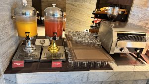 A buffet&#039;s juice counter has two large glass dispensers of mango and papaya juice next to rows of empty glasses and a conveyor toaster.