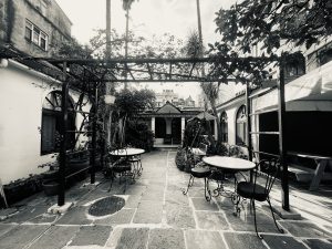 A black and white photo of an outdoor courtyard with a stone-tiled floor. Two small, round tables with wrought-iron chairs sit beneath a vine-covered metal pergola.