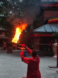 

A person in a red traditional outfit holds a flaming torch in a temple courtyard, with ornate structures, draped red fabric, and smoke-lit surroundings.
