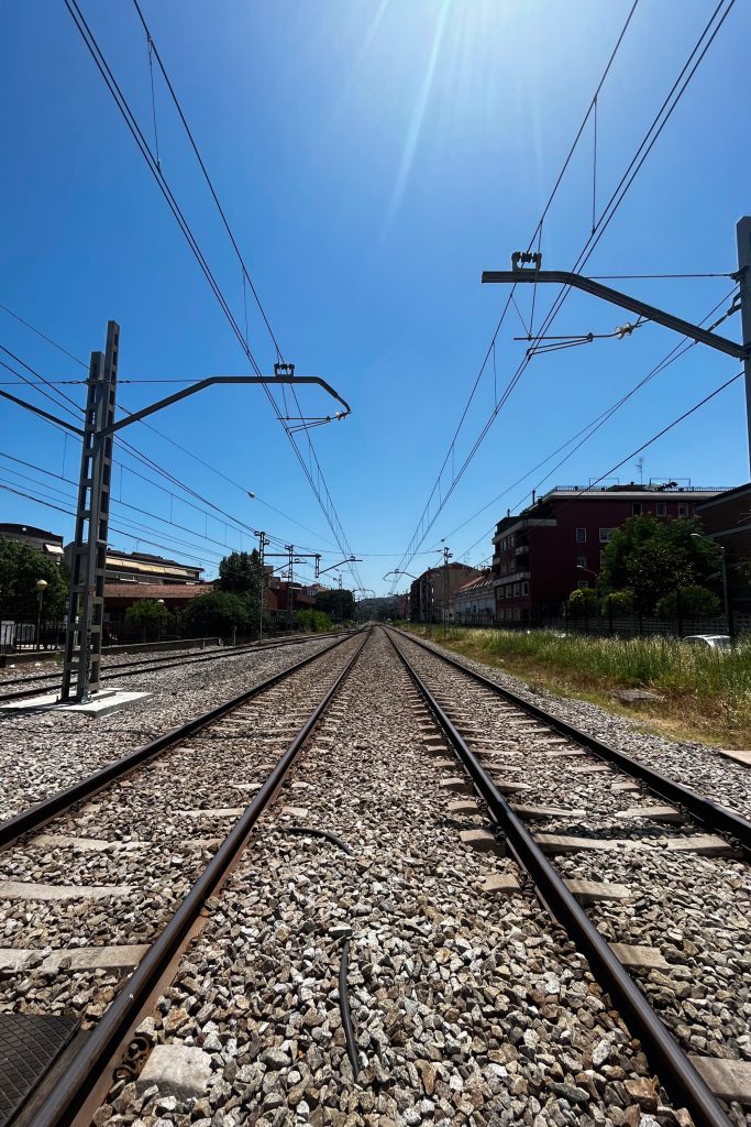 Railway tracks stretch into the distance under a bright blue sky, flanked by grass, gravel, and nearby buildings with overhead wires above.