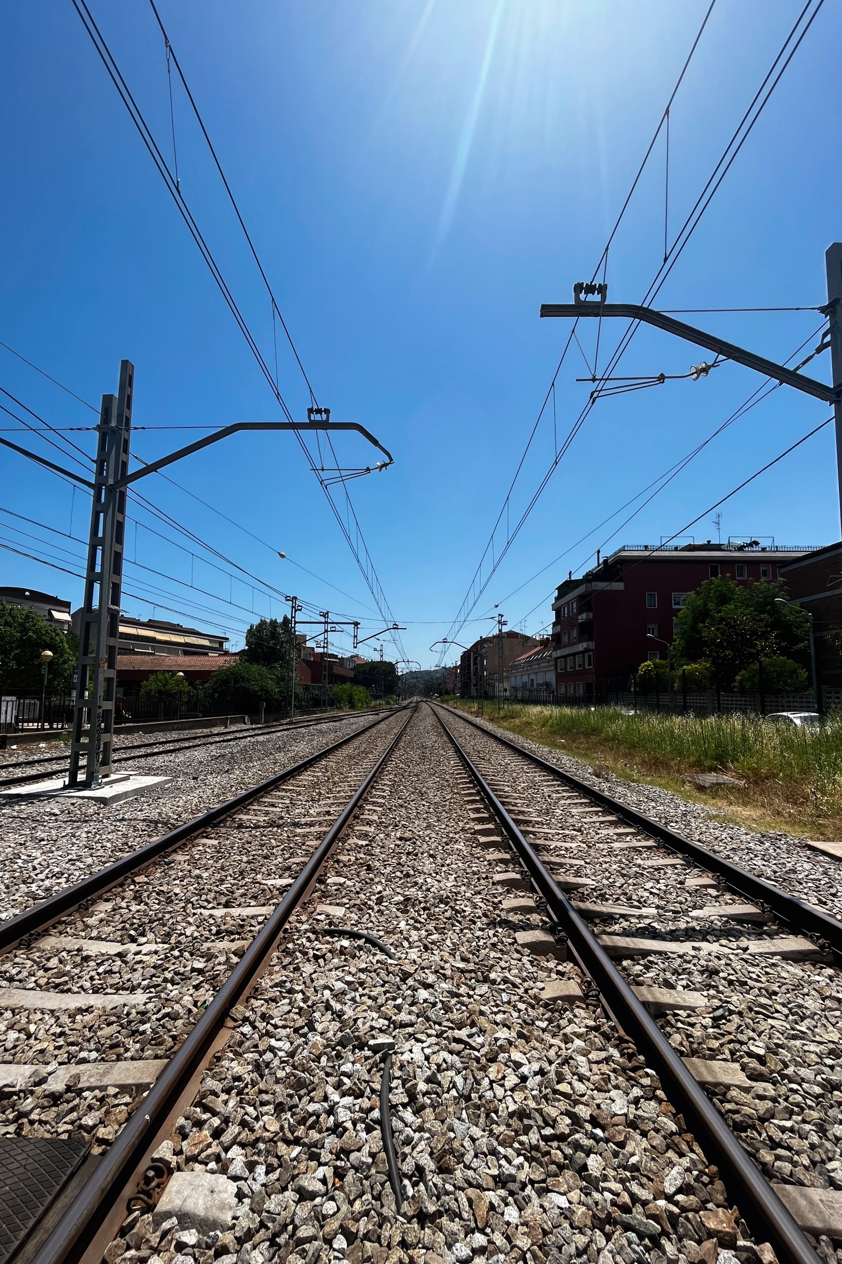 Railway tracks stretch into the distance under a bright blue sky, flanked by grass, gravel, and nearby buildings with overhead wires above.