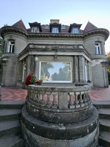A bouquet of red roses lies on a stone railing in front of Pittock Mansion. The large window reflects tall trees nearby, adding depth to the elegant French-style architecture, captured during a calm evening in Portland, Oregon. 