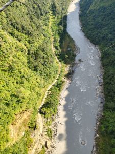 Aerial view of a winding dirt path alongside a river, surrounded by lush green hills and dense trees. 