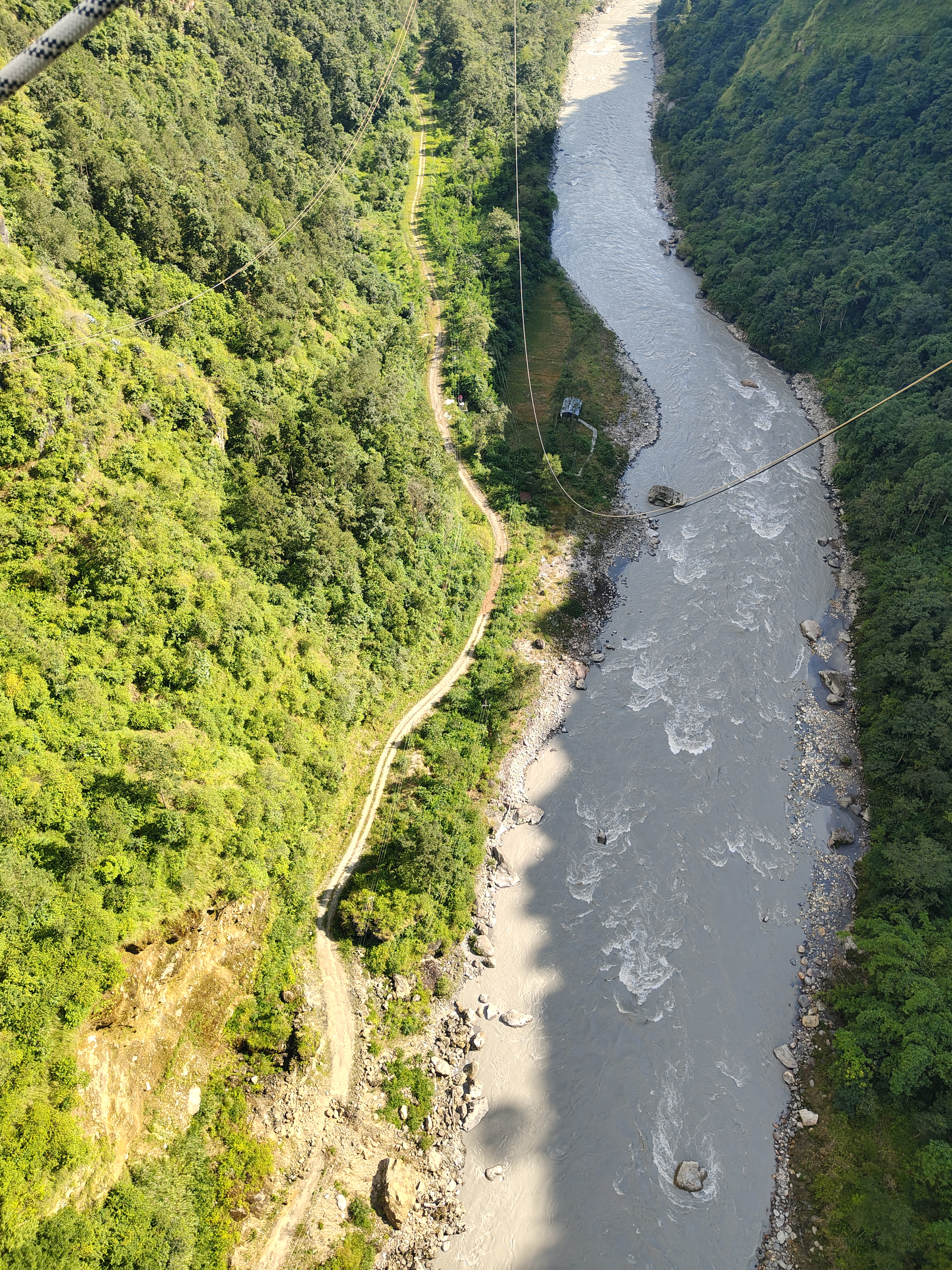 Aerial view of a winding dirt path alongside a river, surrounded by lush green hills and dense trees.