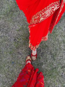 Two female wearing red sarees and red slippers, flaunting their feet, standing on the dried grass ground. One of them is wearing a silver anklet.